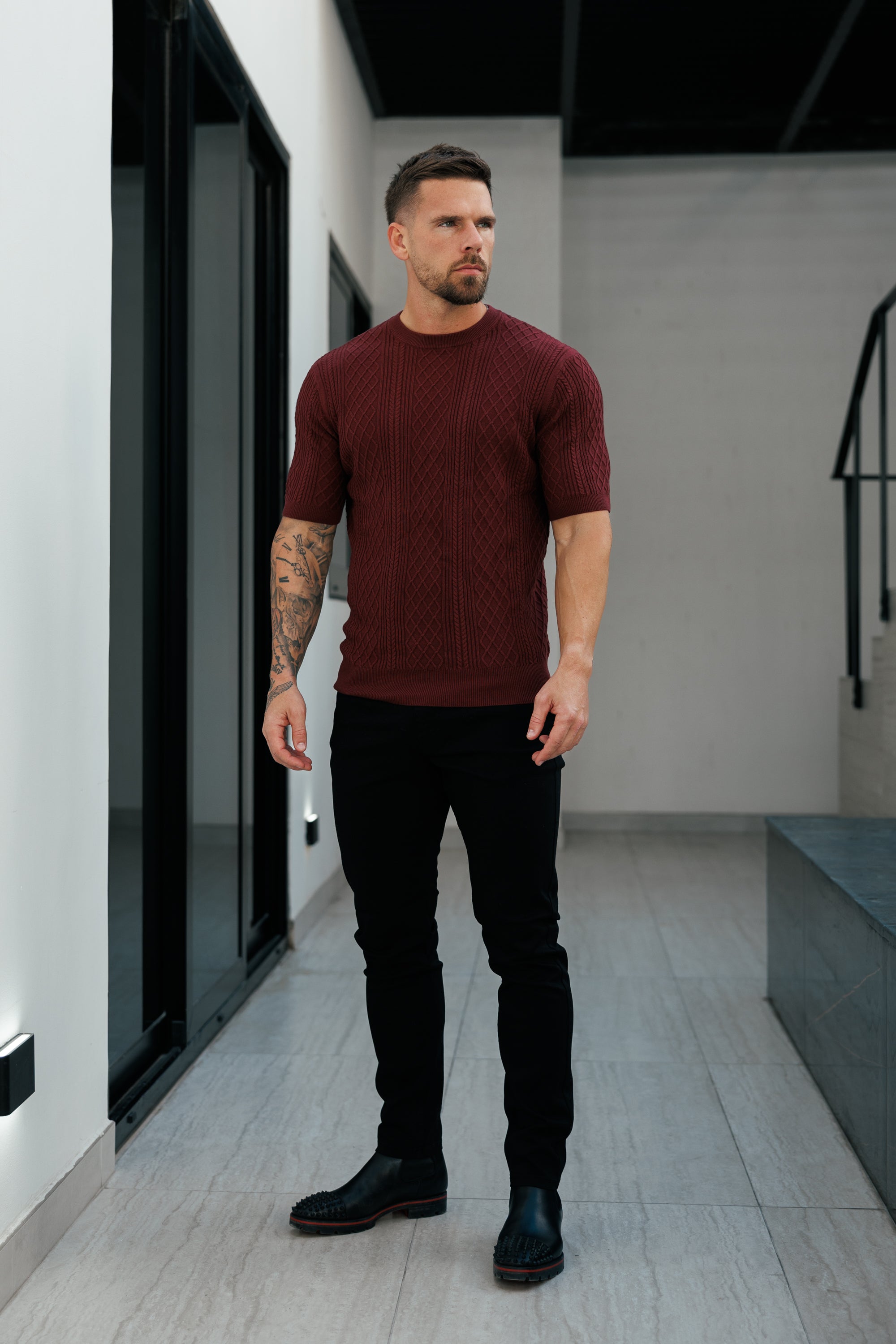 Man wearing a burgundy sweater and black pants standing in a modern indoor setting.